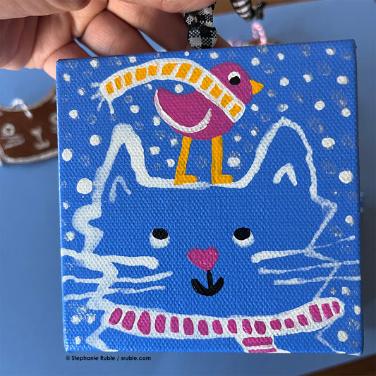 hand holds black and white ribbon attached to small square light blue painting with a white line art cat wearing a pink striped scarf, with a pink heart nose. there's a pink bird with a yellow striped scarf standing on the cat's head and there's snow in the background