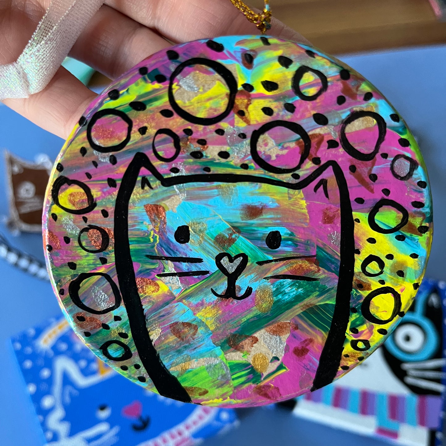 hand holding round ornament with splashes of color. black line art of a sleek cat and circles and dots are on top of the colors