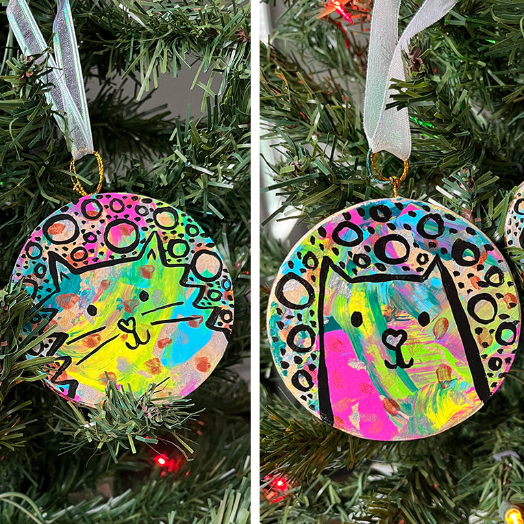 two cropped pictures in one square, with circle cat ornaments hanging on a Christmas tree on each side. ornaments have splashes of colorful paint. black line art and circles and dots on top of colors. left: fluffy cat / right: sleek cat.