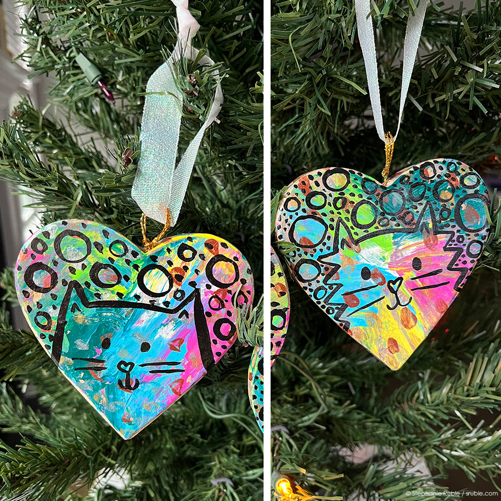 two cropped pictures in one square, with heart cat ornaments hanging on a Christmas tree on each side. ornaments have splashes of colorful paint. black line art and circles and dots on top of colors. left: sleek cat / right: fluffy cat. 