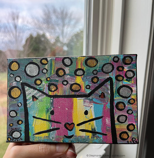 hand holds small horizontal painting of a colorful striped cat face with a heart shaped nose. the background has splashes of rainbow colors and shiny metallic circles in silver and gold. behind the painting is a window showing trees and sky