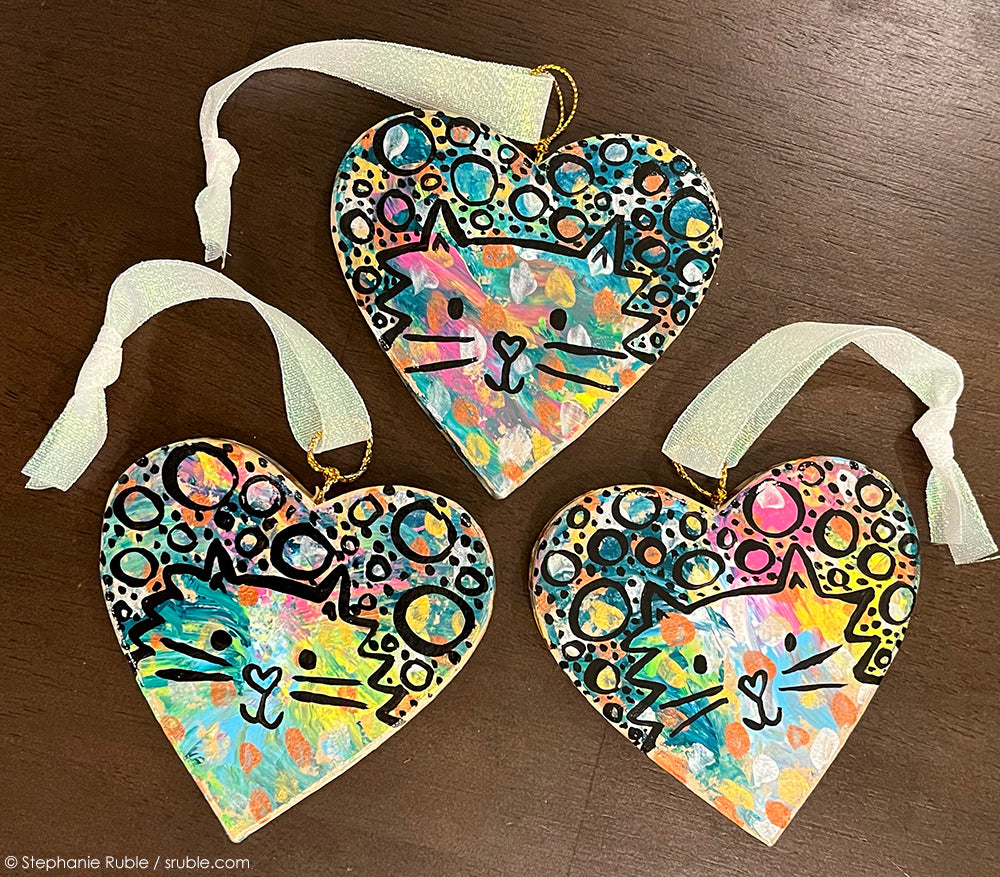 three heart cat ornaments with splashes of colorful paint and fluffy kitties and circles and dots on top with black line. ribbons attached to the top