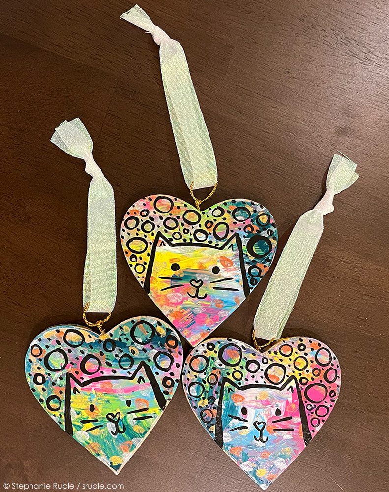 three heart cat ornaments with splashes of colorful paint and sleek kitties and circles and dots on top with black line. ribbons attached to the top