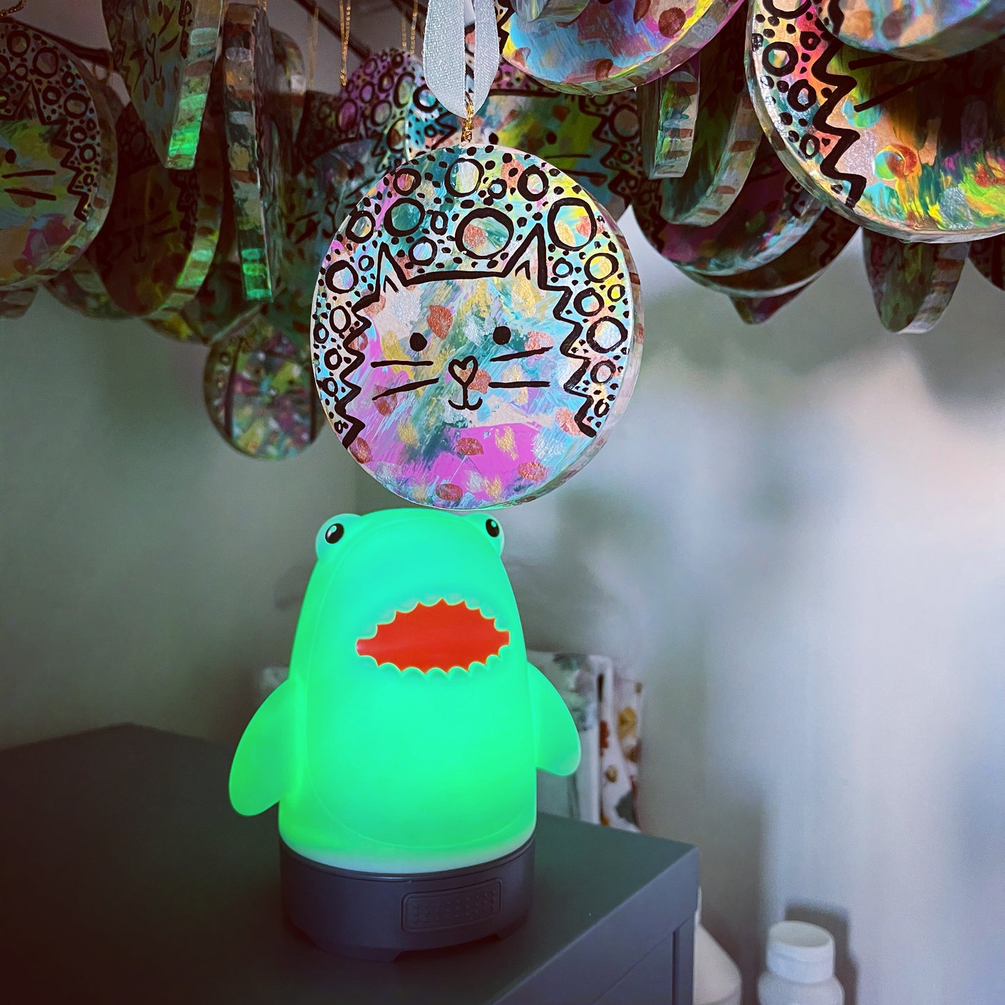 small shark shaped lantern sitting on cabinet looking up at, and with its green light shining on, a circle cat ornament hanging down from above. There are lots of other circle cat ornaments hanging down in the background, but only part of them is showing. ornaments have splashes of colorful paint. black line art of the cat with circles and dots are on top of the colors. 